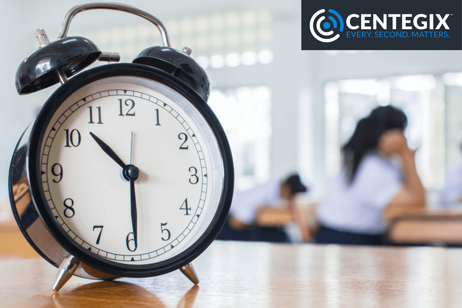 A clock sitting on a teacher's desk with students out of focus in the background.