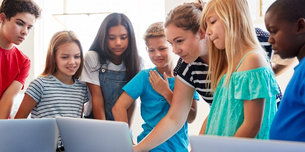 A teacher pointing to a laptop screen surrounded by her young students.